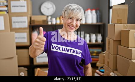 Una donna matura sorridente con i capelli grigi che indossa una t-shirt "volontaria" regala un pollice in un centro donazioni circondato da scatole. Foto Stock
