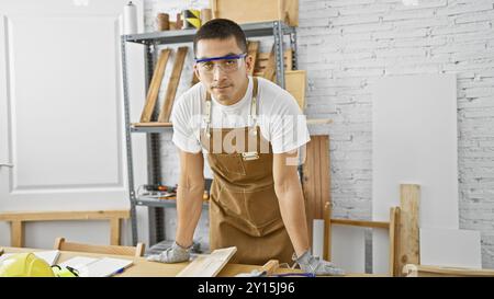 Il bel giovane ispanico che indossa un grembiule si trova in un'officina di falegnameria ben attrezzata. Foto Stock