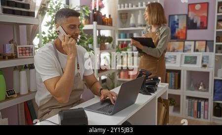 Uomo e donna che lavorano insieme in un negozio di arredamento per la casa con l'uomo durante una telefonata e utilizzando un computer portatile, mentre la donna prende appunti su un appunti, surr Foto Stock