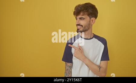 Un bel giovane ispanico con barba e tatuaggi che puntano a lato su uno sfondo giallo isolato Foto Stock