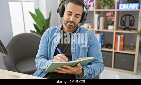 Uomo ispanico con la barba che indossa le cuffie e scrive su un notebook presso lo studio radio Foto Stock