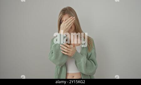 Una giovane donna bionda con un maglione verde copre gli occhi con la mano su uno sfondo bianco Foto Stock