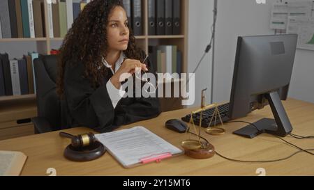 Giovane donna seduta in un ufficio con indosso una veste nera, che contempla mentre tiene gli occhiali, circondata da documenti legali, un martelletto e bilance di giusto Foto Stock