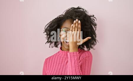 Felice donna africana con capelli ricci che indossa occhiali e maglione rosa che copre un occhio con la mano su uno sfondo rosa Foto Stock