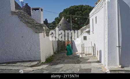 Una giovane donna ispanica in un abito verde cammina lungo una strada affascinante nella storica città di alberobello, in italia, circondata da iconici trulli che ospitano un Foto Stock