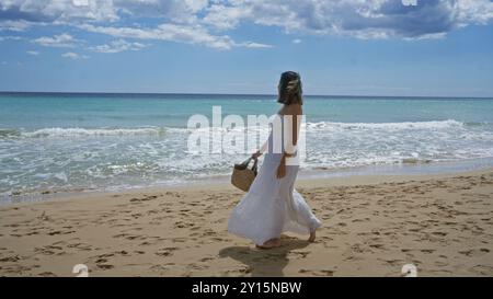 Una bella giovane donna ispanica passeggia lungo la splendida spiaggia di pescoluse in puglia, italia, portando con sé una borsa intrecciata e indossando una dres bianca fluente Foto Stock