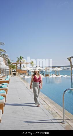 Una donna elegante cammina accanto a una lussuosa piscina a sfioro in un resort, incarnando svago ed eleganza. Foto Stock
