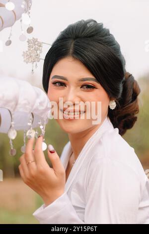 Affascinante giovane donna indonesiana sorridente davanti alla macchina fotografica, indossa abiti tradizionali giavanesi moderni, kebaya nuziale in velluto bianco in una b all'aperto Foto Stock