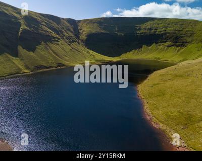 La luce del sole si riflette su un lago blu annidato in una valle circondata da montagne con erba verde in una giornata di sole Foto Stock