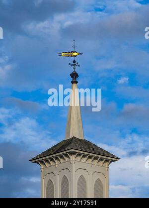 Un limp giallo si libra graziosamente sopra un alto campanile bianco della chiesa, con uno sfondo di cielo blu e nuvole sparse, catturando una serena atmosfera pomeridiana. Foto Stock