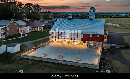 Ronks, Pennsylvania, USA, 19 luglio 2024 - tranquilla fattoria al tramonto con Un grande fienile rosso con Un Silo bianco, un cortile in pietra illuminato, Una vicina Brick House e ampi campi verdi sotto Uno splendido cielo al tramonto, creando Un ambiente tranquillo e pittoresco. Foto Stock