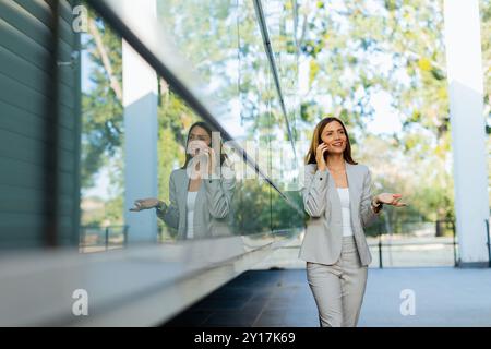 Una donna professionista cammina con facilità, sorridendo mentre si concentra sul telefono, mescolando lo stile di vita moderno con la tranquillità della natura sullo sfondo Foto Stock