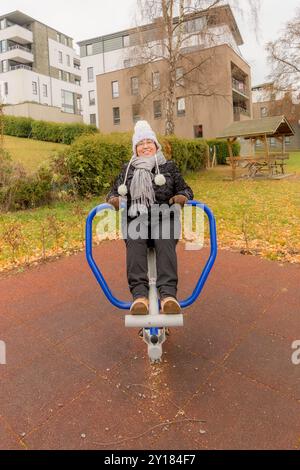 Donna di mezza età in palestra all'aperto nel parco pubblico, che fa esercizi alle gambe, indossa abbigliamento invernale, giacca, cappello e guanti su nuvoloso da Foto Stock