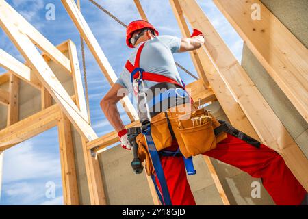 Un operaio edile con elmetto rosso e cintura portautensili è impegnato a fissare travi in legno in una struttura parzialmente costruita sotto un cielo azzurro. Foto Stock