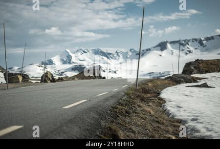 Una tortuosa strada di montagna norvegese si estende in lontananza, fiancheggiata da cime innevate e macchie di neve lungo la strada sotto un blu chiaro Foto Stock