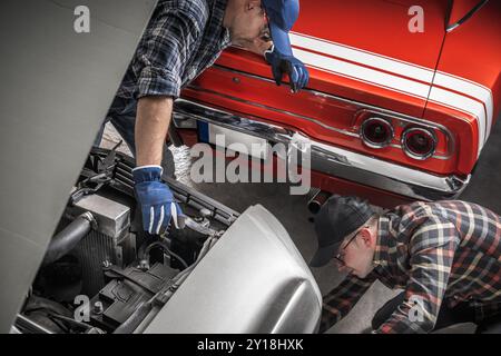 Due meccanici si concentrano sulla riparazione di un'auto d'epoca in un garage, mentre un'altra auto d'epoca è parcheggiata nelle vicinanze, mostrando il loro lavoro di squadra e la loro abilità. Foto Stock