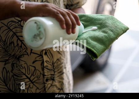 Una persona tiene una bottiglia di soluzione detergente su un panno verde, preparandosi a pulire una superficie all'aperto, circondata da un pavimento piastrellato e luce naturale. Foto Stock