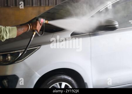 Una persona spruzza acqua su un'auto argentata utilizzando un tubo flessibile ad alta pressione in un garage. L'azione mostra attenzione ai dettagli durante il lavaggio del veicolo. Foto Stock