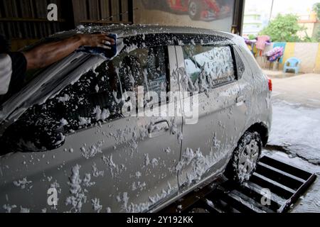 Una mano sfrega l'esterno di un'auto color argento ricoperta di schiuma in un autolavaggio, con uno sfondo urbano caratterizzato da opere d'arte colorate e un'area salotto. Foto Stock