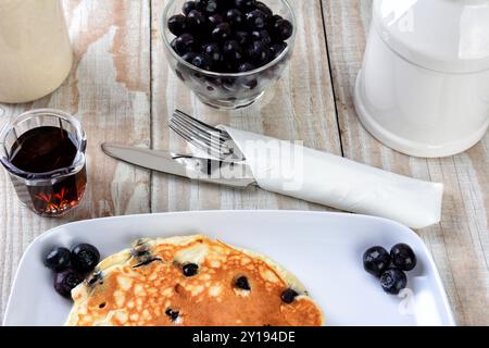 Primo piano di una pila di pancake di mirtilli freschi fatti in casa su un piatto bianco. Utensili e stoviglie per sciroppo d'acero e un tavolo in legno rustico. Foto Stock