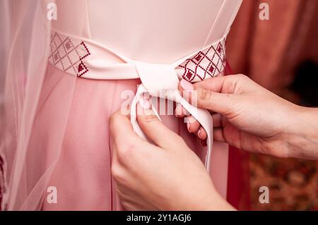 Elegante primo piano di mani che lega un fiocco a un abito rosa con ricami intricati. Foto Stock