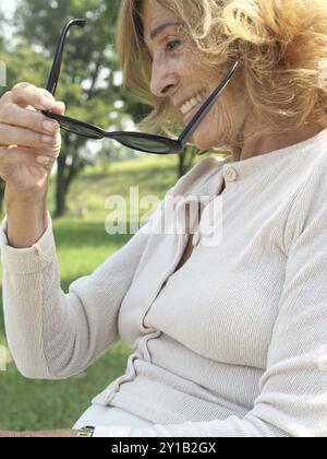 Una donna anziana serena giace in un parco tranquillo, immerso in una lussureggiante erba verde. Crogiolati al caldo sole estivo. Tenendo i suoi occhiali da sole con un sorriso. Embodyin Foto Stock