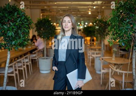 Una donna sicura di sé in costume da lavoro con un laptop in mano in piedi nel ristorante della hall guarda la fotocamera Foto Stock