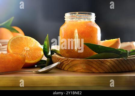 Dettaglio del vaso con marmellata d'arancia e tagliare la frutta intorno ad esso su banco da cucina in legno e sfondo scuro. Vista frontale. Foto Stock