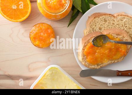 Dettaglio della marmellata d'arancia spalmata su una fetta di pane e in contenitori di vetro su una tavola di legno con burro e posate. Vista dall'alto. Foto Stock