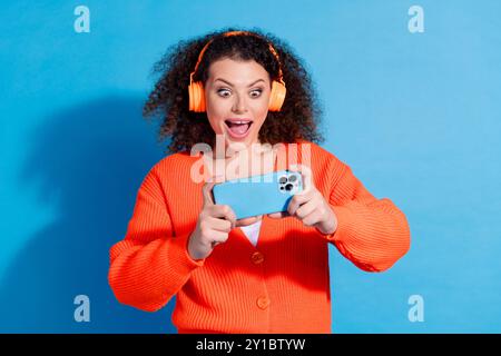 Foto ritratto di una modella dai capelli ondulati che indossa un cardigan arancione che gioca a un gioco mobile isolato su sfondo blu Foto Stock