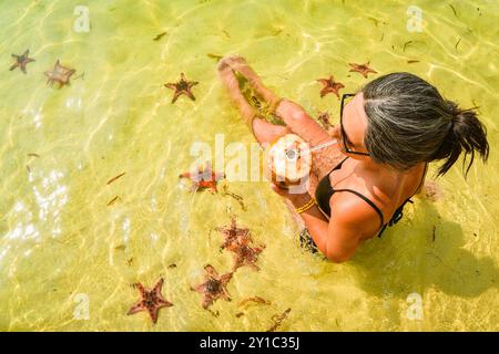 Una donna felice in bikini sulla spiaggia di stelle marine beve una bevanda fresca al latte di cocco sulla spiaggia di pesce stellato godendoti la famosa isola tropicale di Koh Rong, Cambogia. Foto Stock