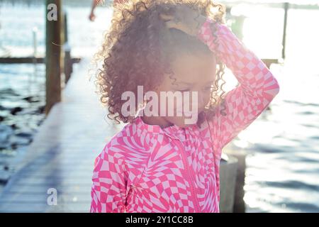 Ragazza sorridente in posa rosa per molo, luce del sole che si riflette sull'acqua Foto Stock