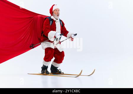 Babbo Natale sciando, tirando dietro di sé un grande mantello rosso, afferrando pali, espressione concentrata, su sfondo bianco studio. Foto Stock