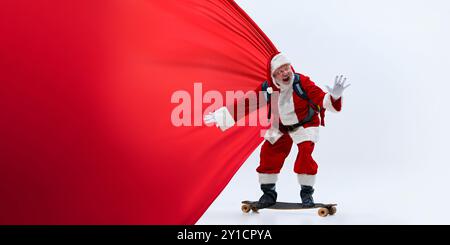 Babbo Natale in sella allo skateboard tenendo in mano un grande mantello rosso, con un'espressione entusiasta su sfondo bianco studio. Foto Stock