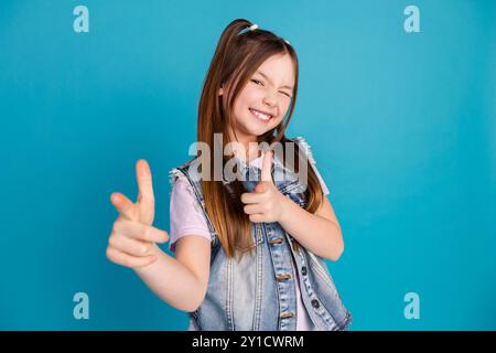 Ritratto fotografico di una bambina carina che ti indica allegri abiti in denim eleganti vestiti con l'occhiolino, isolati su sfondo blu Foto Stock