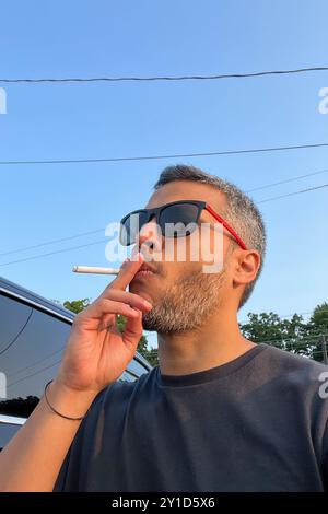 Un uomo sta fumando una sigaretta mentre indossa gli occhiali da sole. Concetto di relax e svago, mentre l'uomo si prende una pausa dalla sua giornata per gustare un sigaretto Foto Stock