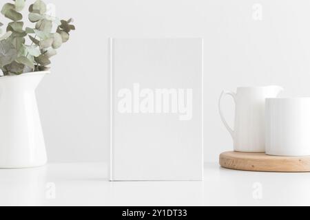 Modello di libro bianco con eucalipto in una pentola e una tazza su un tavolo bianco. Foto Stock