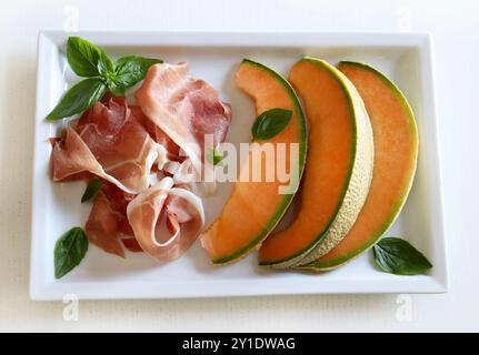 Prosciutto e melone decorati con foglie di basilico fresche isolate su sfondo bianco. Piatto estivo. Direttamente sopra. Foto Stock