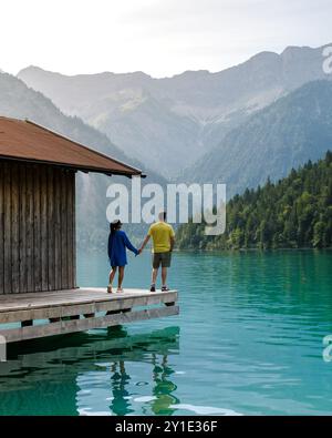 Una coppia sta mano nella mano su una piattaforma di legno vicino alle limpide acque blu di Plansee Austria, circondato da maestose montagne e lussureggiante vegetazione unde Foto Stock