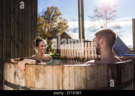 Giovane coppia che si diverte nella vasca idromassaggio all'aperto, immersa nell'acqua calda, circondata dalla natura incontaminata, da vicino. Glamping e concetto di relax. Foto Stock