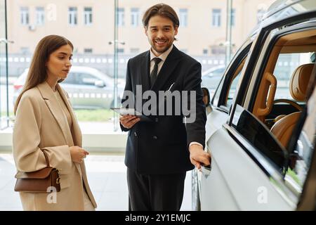 Immagine completa media di un concessionario di auto sorridente maschio in tuta nera che apre la portiera dell'auto, presentando il modello dimostrativo a una cliente donna che intende acquistare un nuovo veicolo presso il centro della concessionaria Foto Stock