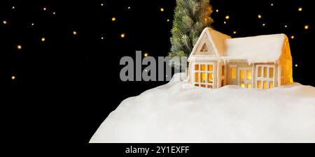 Gingerbread House Day. Biglietto di Natale. Casa con illuminazione calda e accogliente all'interno. Una casa su una collina, porte coperte di neve, tetto innevato. Stelle, fiocchi di neve Foto Stock