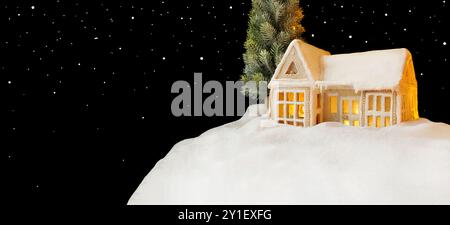 Gingerbread House Day. Biglietto di Natale. Casa con illuminazione calda e accogliente all'interno. Una casa su una collina, porte coperte di neve, tetto innevato. Stelle, fiocchi di neve Foto Stock