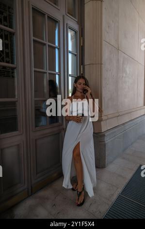 Una donna con un vestito bianco si trova accanto a una porta di vetro, tenendo un telefono e una tazza. Guarda il telefono mentre si appoggia contro il muro. Foto Stock