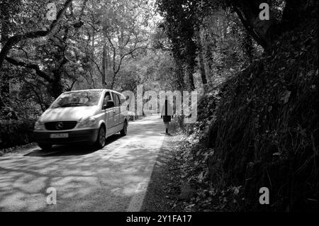 Monocromatico di una donna che cammina lungo una stretta strada forestale mentre passa un minivan a Sintra Foto Stock