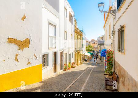 Una pittoresca strada collinare con caffetterie e negozi sul marciapiede nella cittadina di mare imbiancata di Lagos, sulla costa dell'Algarve del Portogallo. Foto Stock