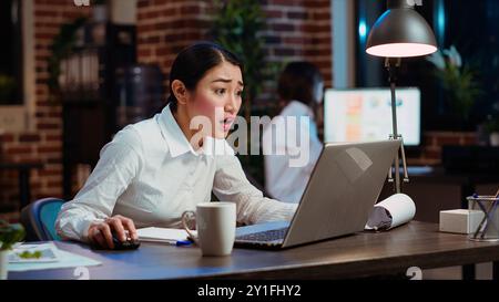 I dipendenti asiatici sono sorpresi, scioccati dalla risposta negativa ricevuta dai clienti mentre discutono di questioni aziendali tramite e-mail. Il lavoratore sta brillando mentre guarda lo schermo del notebook, sentendosi deluso Foto Stock