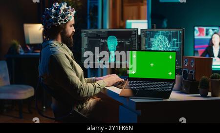 Ingegnere con auricolare EEG per la programmazione del trasferimento cerebrale nel mondo virtuale del computer accanto al notebook con schermo verde. Transumanista che usa la neuroscienza per ottenere l'anima digitale, il dispositivo di mockup, la telecamera A. Foto Stock