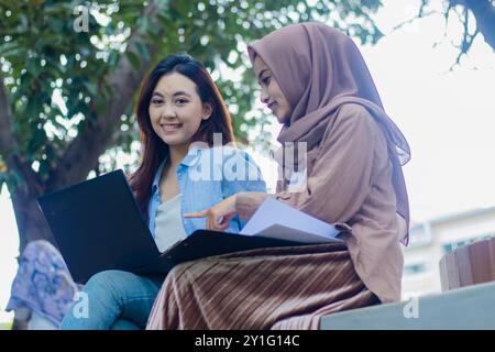 Un paio di bellissime ragazze asiatiche che guardano la telecamera stanno discutendo insieme di portare laptop, libri e appunti mentre puntano nel parco del campus. Foto Stock