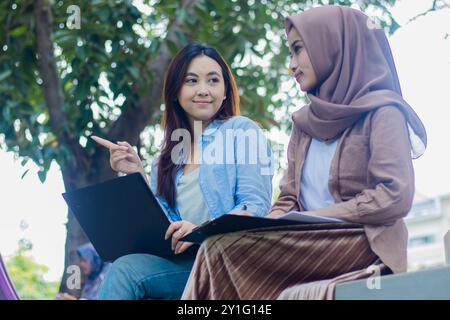 un paio di graziose donne indonesiane mostrano con franchezza qualcosa che rimane quando discutono del compito di portare un portatile, libri e appunti in cam Foto Stock